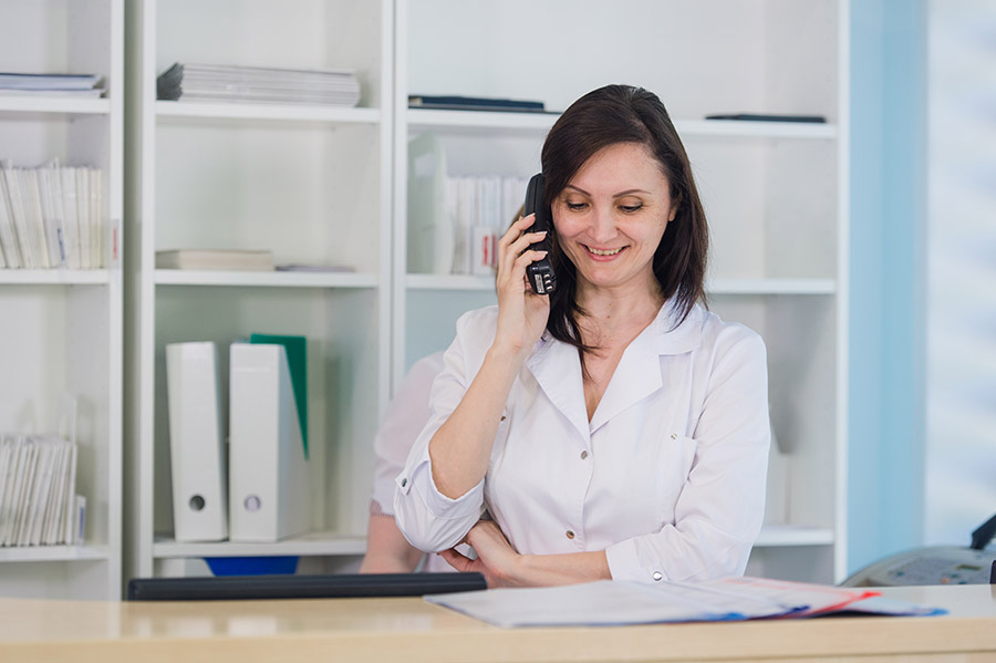 Young practitioner doctor working at the clinic reception desk, she is answering phone calls and scheduling appointments